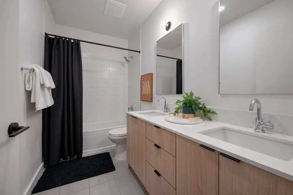 A modern bathroom featuring dual sinks, a large mirror, sleek cabinetry, a spacious shower area, and a touch of greenery with a potted plant. The room feels fresh, inviting, and thoughtfully appointed for guest comfort.