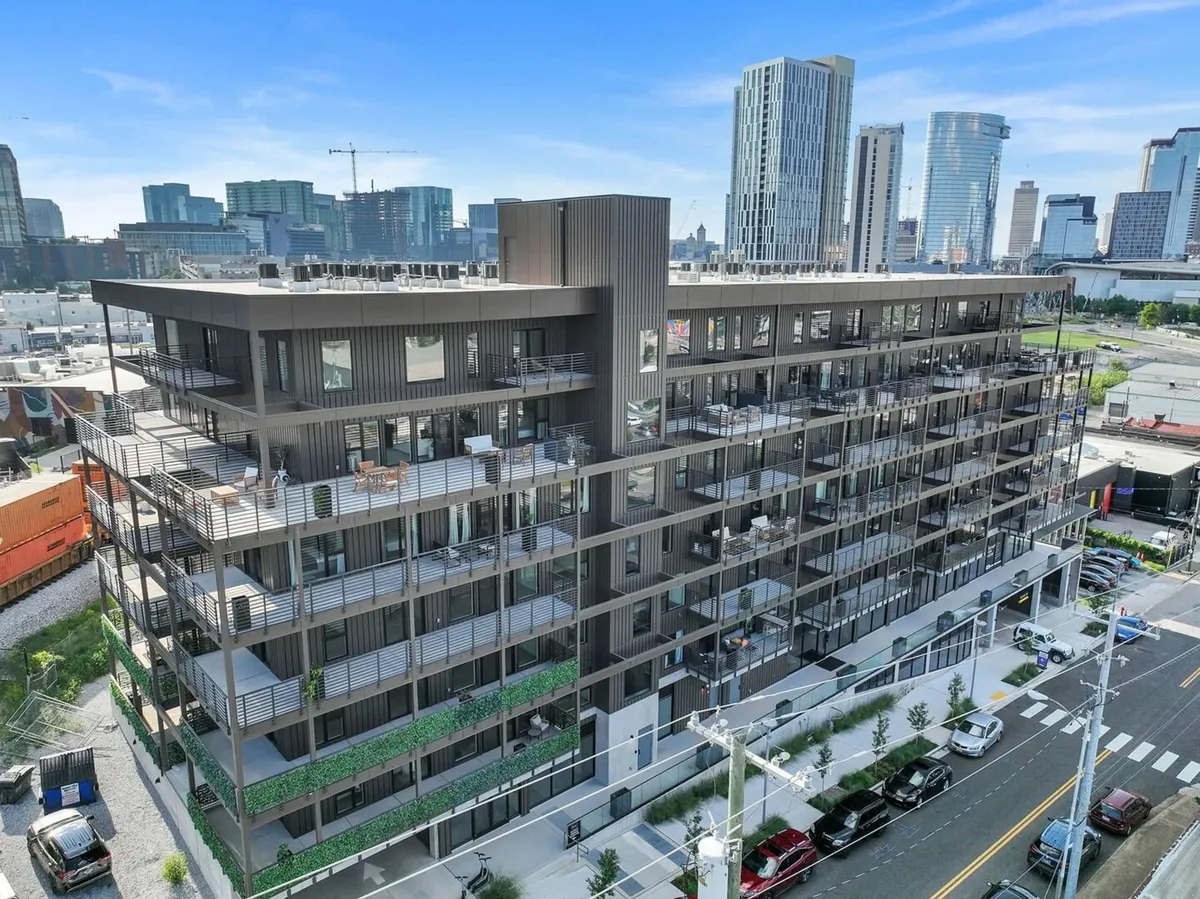 This exterior image highlights the building’s modern architecture, with balconies adorned with greenery and an urban landscape filled with city energy. The city skyline is visible in the background, suggesting a vibrant, creative neighborhood.