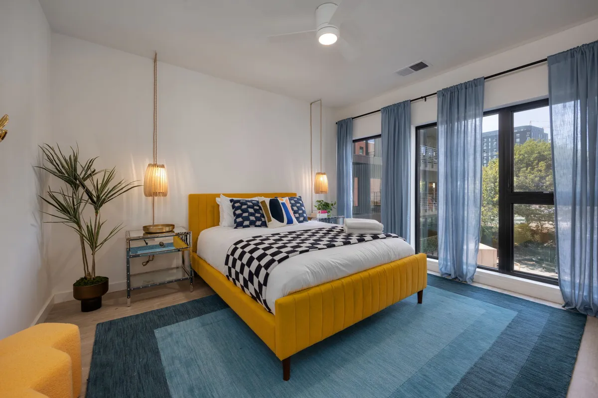 A cozy bedroom with a yellow bed adorned in checkerboard bedding, a blue rug, large windows, and statement pendant lights. The room looks cheerful and modern, embodying both comfort and playfulness.