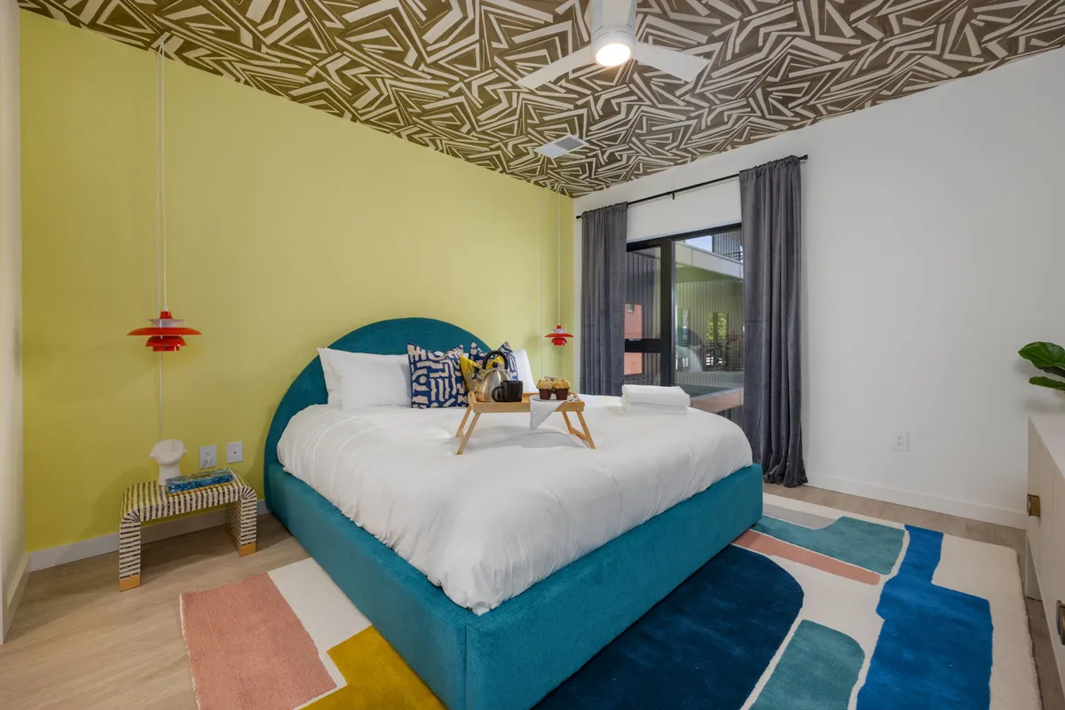 A cheerful bedroom featuring a teal headboard, colorful area rug, decorative ceiling, stylish bedside table, and a window with curtains. The space is full of natural light and playful decor touches, inviting relaxation and comfort.