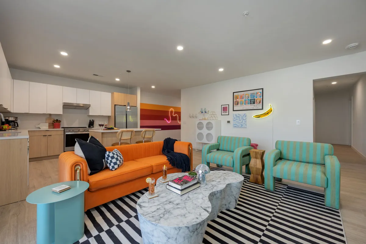 A vibrant living space featuring a bold orange sofa, a modern striped rug, a chic marble coffee table, striking colorful wall art, and a playful neon banana light. The room invites guests to relax, socialize, and celebrate in a lively, art-inspired setting.