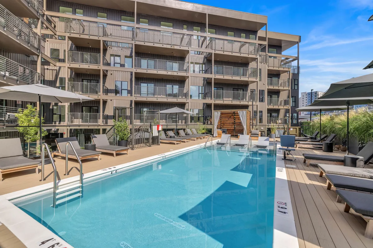 A resort-style outdoor swimming pool surrounded by lounge chairs, a wooden deck, lush greenery, and the contemporary building facade. The scene is tranquil, with hints of group relaxation and socializing by the water.