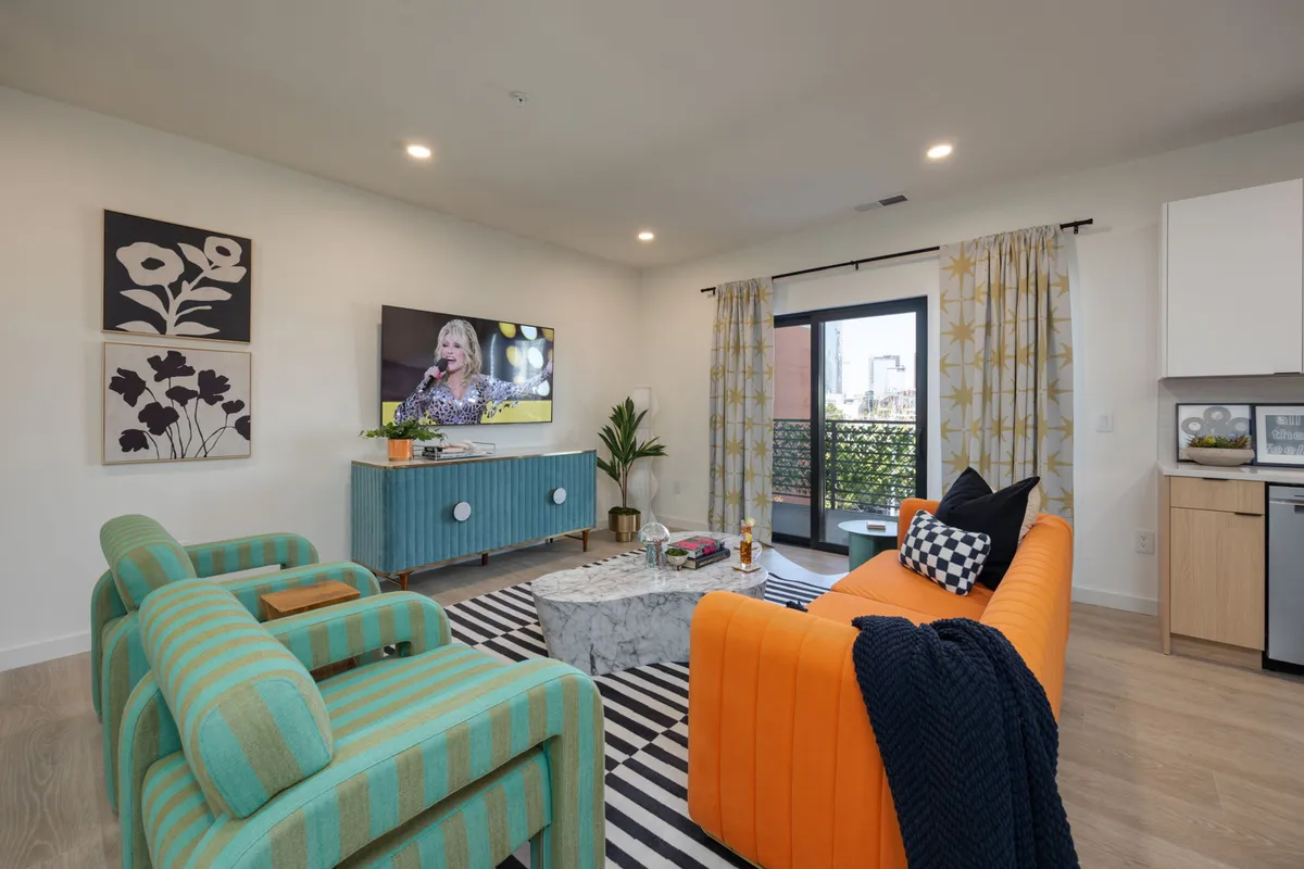 A vibrant living area with a bold orange sofa, striped black-and-white rug, a large TV showing a performance, and colorful modern art on the wall. The setup is open, social, and full of energy, inviting guests to gather and unwind.
