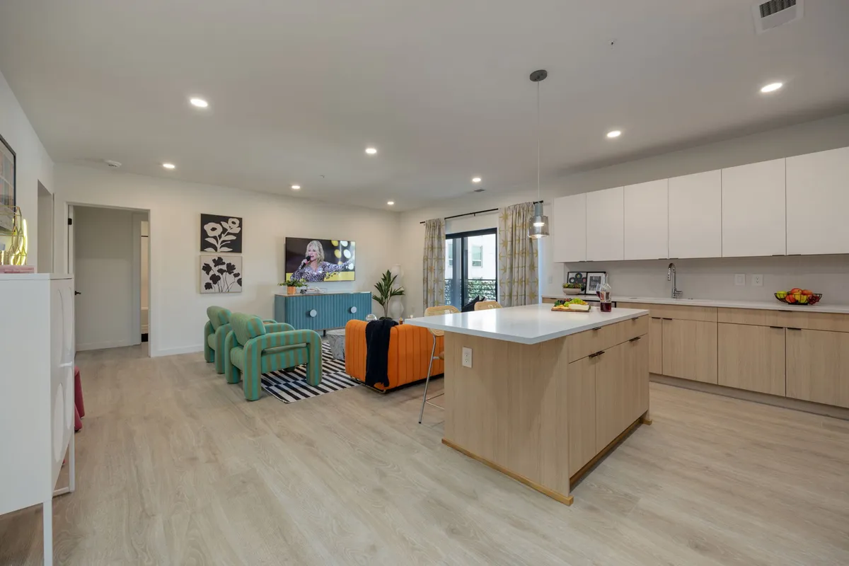 A modern, cheerful interior scene with green armchairs, an orange sofa, a kitchen island, a television, and decorative plants. The area is set up for relaxation, entertainment, and group gatherings, creating a sense of homey comfort and urban style.