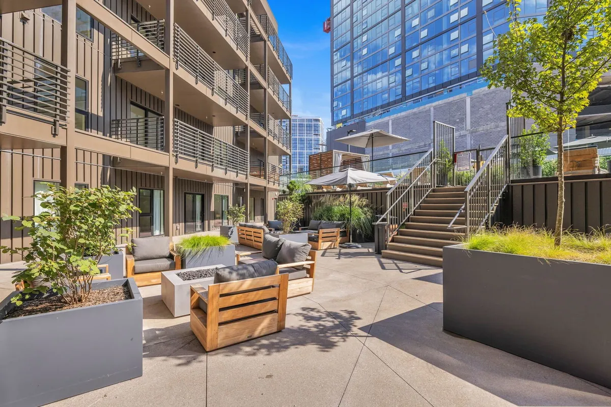 An inviting exterior seating area with comfortable modern furniture, lush planters, and a staircase leading upward. Ideal for social gatherings, outdoor dining, or simply soaking in the sunset in a vibrant setting.